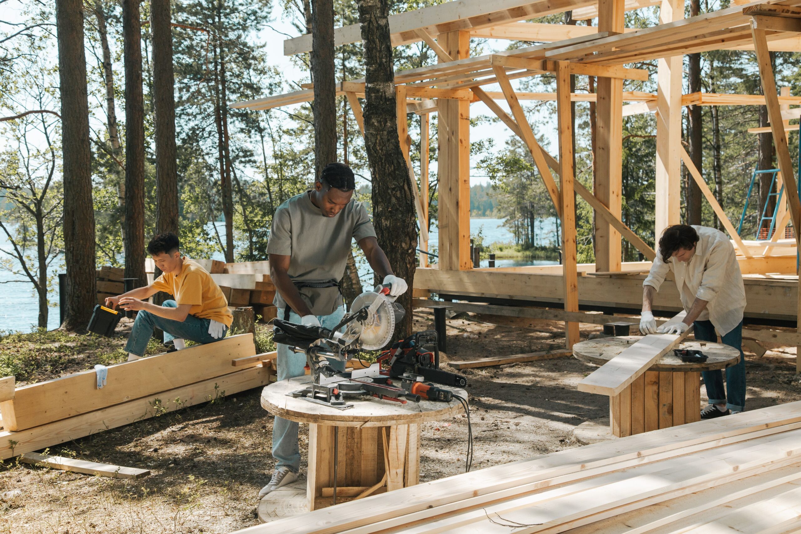 Team of diverse workers engaged in carpentry at a forested lakeside construction site.